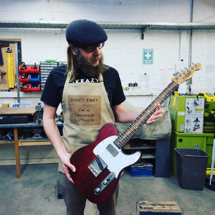 A photo of me in the workshop holding a finished guitar wearing an apron that has printed upon it 'Don't Fret - I'm a Trained Luthier'