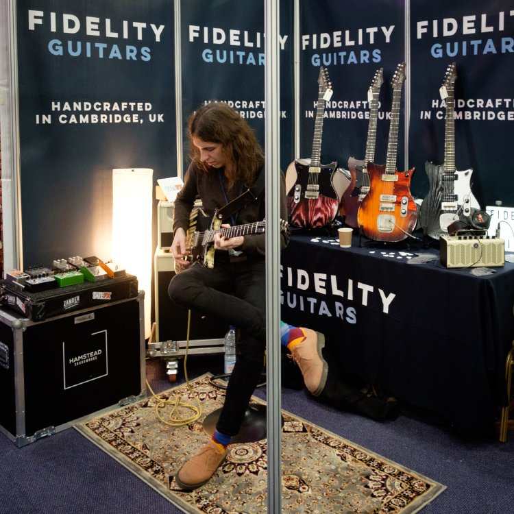 A photo of someone playing a guitar at the Fidelity Guitars booth at a show. Behind them stand 4 more guitars by Fidelity.