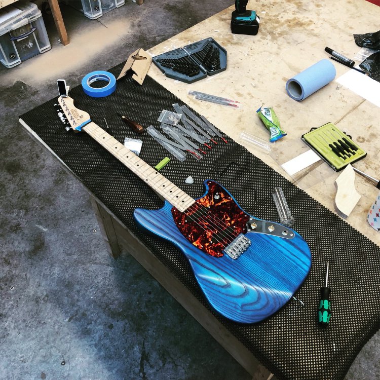 A striking blue electric guitar with a red tort pickguard and maple neck sits on the workbench as it undergoes final tweaking.