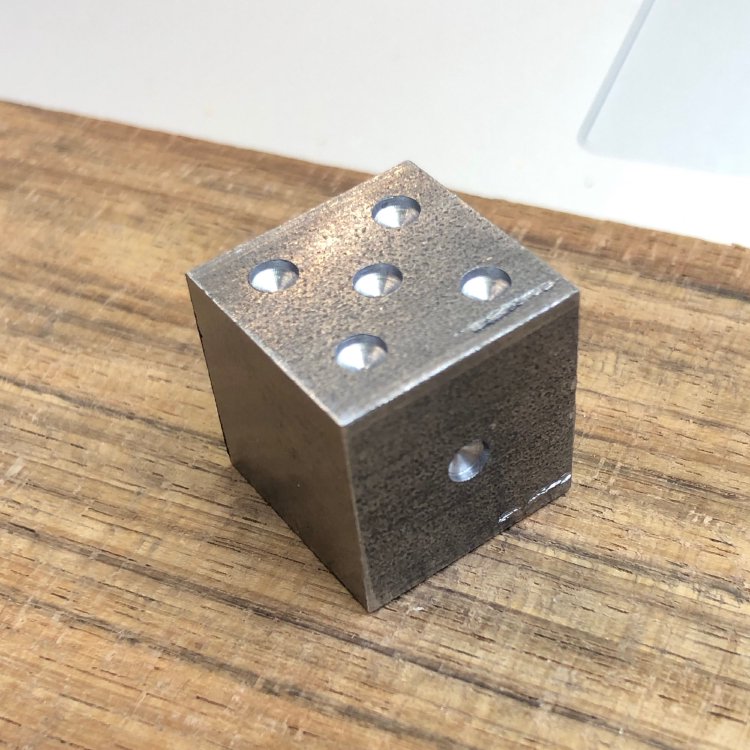 A small metal cube with shallow holes drilled into the sides to make it look like a dice.
