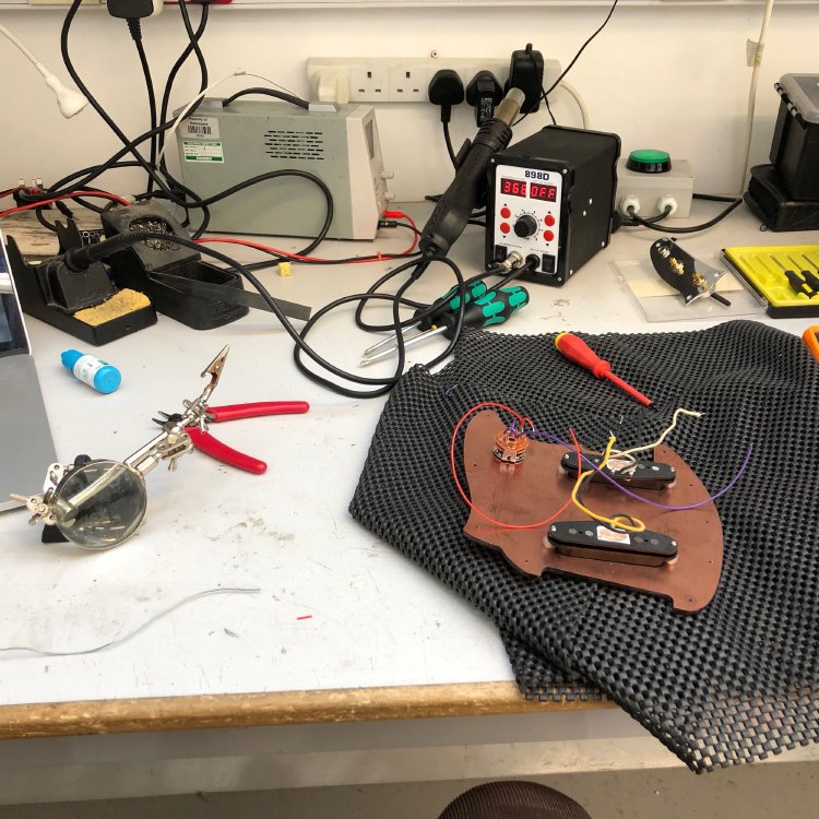A pickguard loaded with pickups and a selector switch is being soldered up on an electronics workbench.