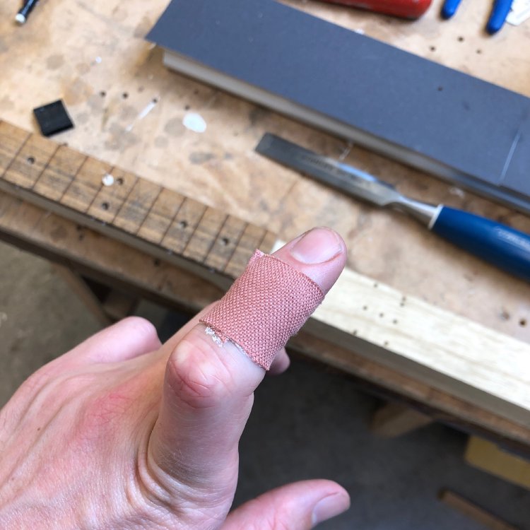 A picture of the author's hand, with a plaster on one finger. In the background we see a workbench with a part made guitar on it, and a chisel beside it.