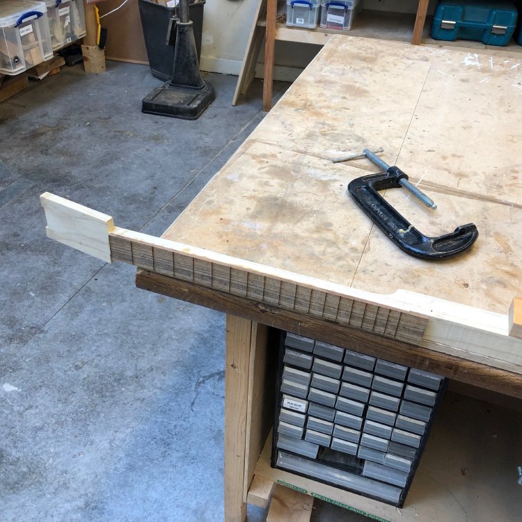 The neck of the cigar box guitar, complete with fretboard and unshaped headstock, is sitting on the edge of the workbench, with a G clamp next to it.