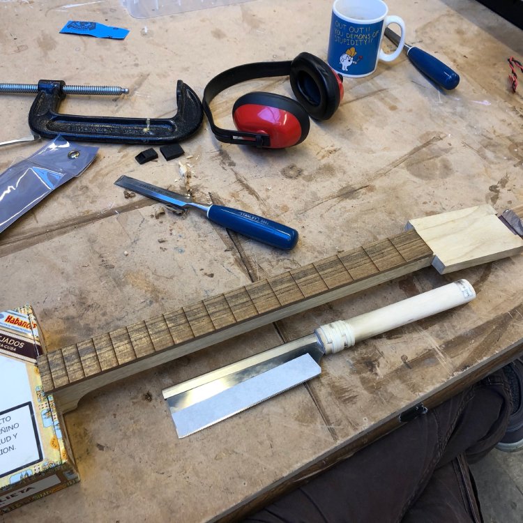 The cigar-box in progress guitar sits on a workbench, showing the fret slots on the neck have been cut. Beside it sits a japanese pull-saw.