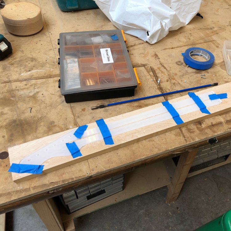 A plank of birds-eye maple sits on the workbench, and taped to it is a laser-cut acrylic template with the outline of a guitar neck.