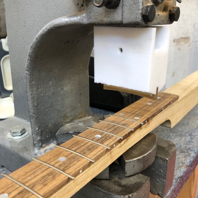The neck sits in an arbour-press, and on one side of the press you can see the fret-slots have been filled with fret wire, and on the other side they have not yet been filled.