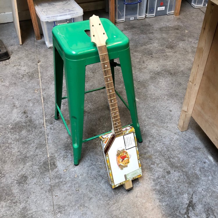 The in-progress cigar box guitar leans against a stool in the workshop. You can see that the headstock now has a more traditional guitar headstock shape to it, and holes for the tuners.