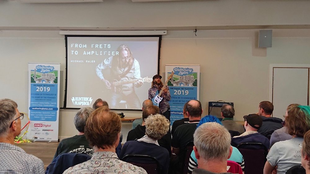 I am presenting to a crowded room, showing the audience somethinb about the guitar I'm holding, in front of a slide titled 'From Frets to Amplifiers'