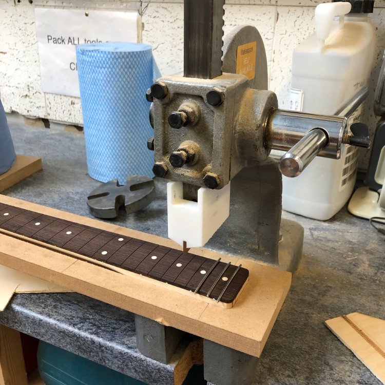 A view of a neck under an arbour-press. The neck has three frets inserted so far, the rest of the fret-slots remain to be filled. On the arbour press is a blocky 3D-printed jig that holds a brass insert that is the fret press.