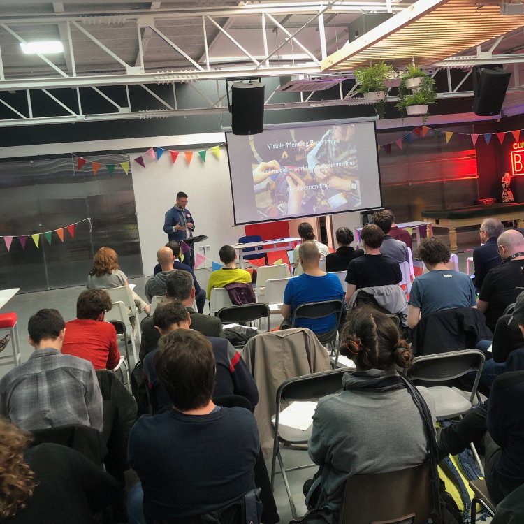 A photo of a speaker presenting slides in front of an audience in a well lit room that has bunting around it.