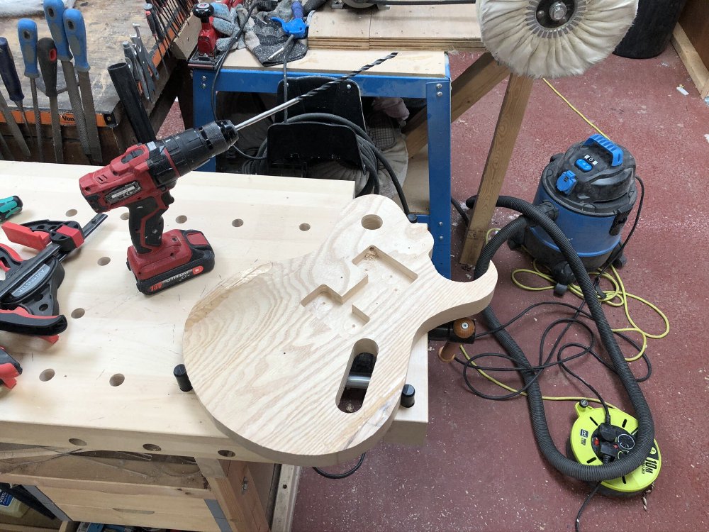 A guitar body sits on the workbench next to a power-drill that has an exceptionally long bit in it (about a foot long).