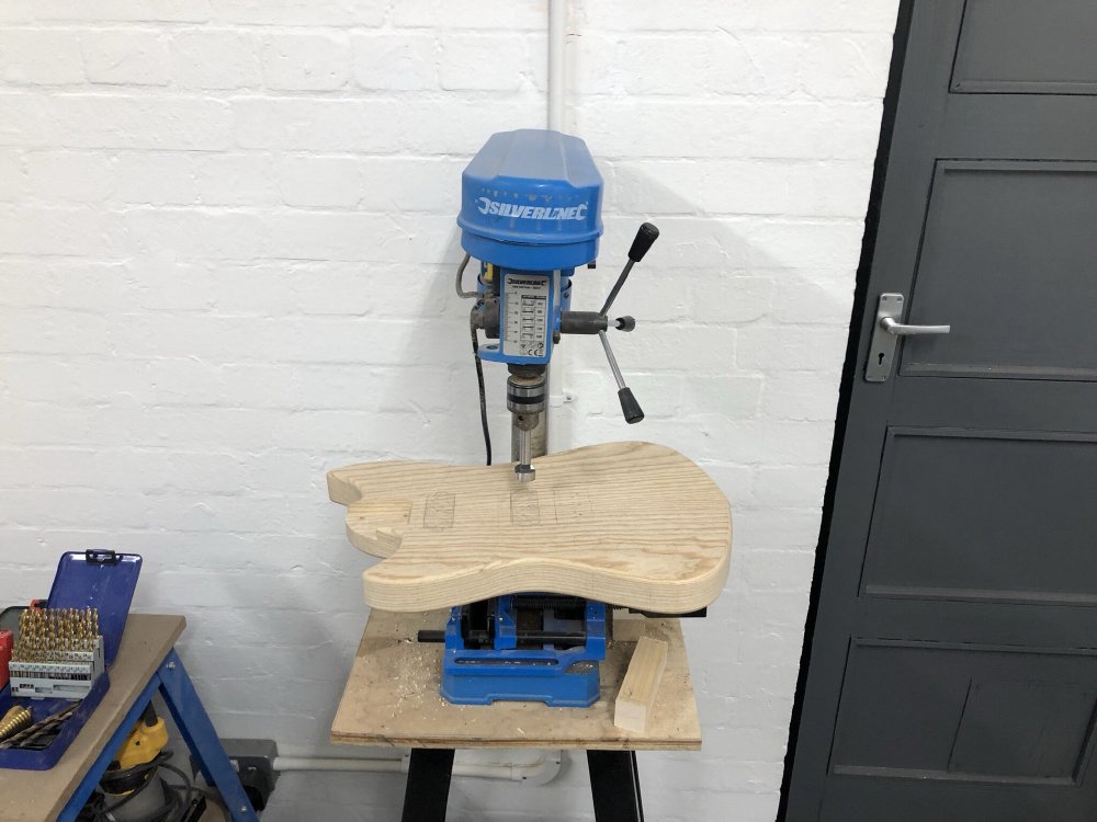 The guitar body sits on the platform of a table-top pillar drill. There is a broad forstner bit in the drill, and on the body you can see the pencil marks for where the pickup cavities will be.
