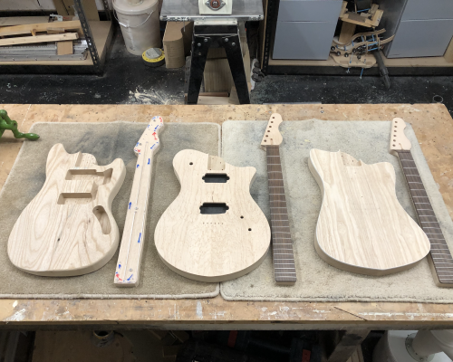 Three work in progress guitars sit on a workbench, each a body with a neck beside it. All are raw wood and unfinished. The left body has pickup cavities and a neck pocket cut, and the neck is just roughly shaped with a template on top. The middle one is the most complete, with the neck fretted and all the body holes cut. The right body has only a neck pocket but no other holes init, but the neck is fretted and otherwise complete.