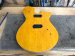 A guitar body sits on the workbench. It has a maple cap, and is stained a deep yellow colour.