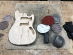 A shaped but unpainted guitar body sits on a workbench surrounded by sanding disk.