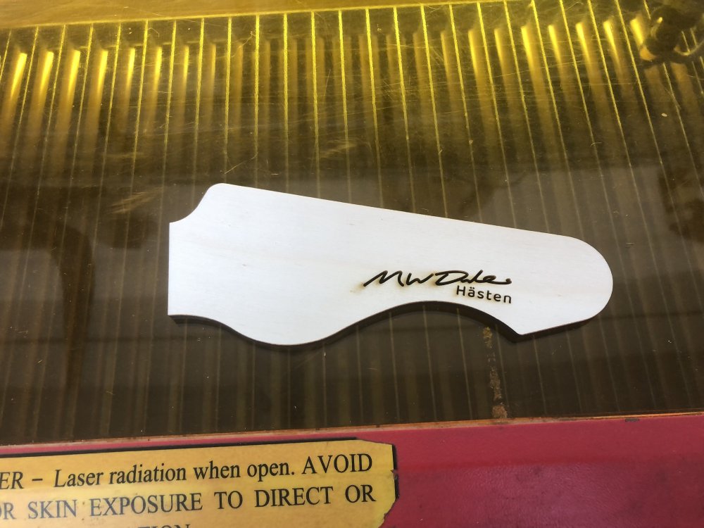 A piece of laser cut ply-wood in the shape of a guitar headstock with 'M W Dales' etched into it in a hand-written scrawl, and then 'Hasten' in an san-serif font.