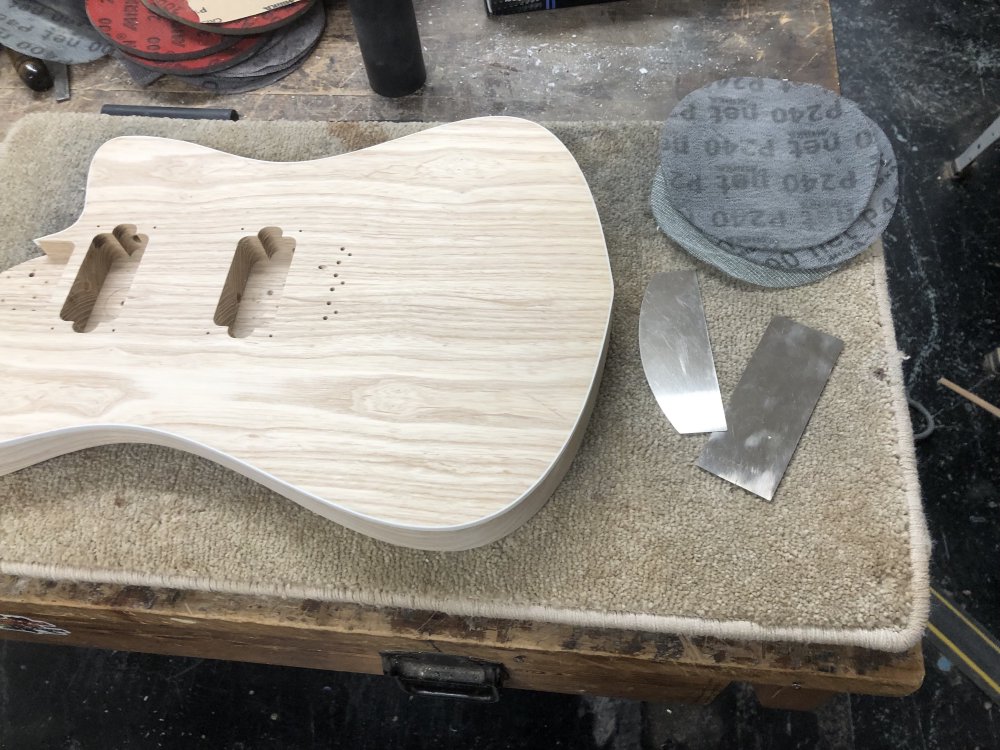 A photo of a guitar body sat on the workbench on a carpet tile, surrounded by metal wood scrapers and sanding disks.