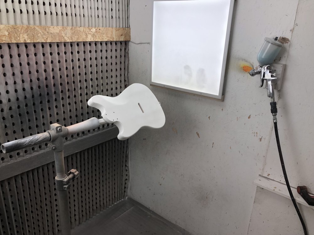A photo of the inside of the spray-booth, with the Mustang-style guitar body mounted on a stand by a stcik mounted in the neck-pocket of the body, to make it easier to spray. On the wall is mounted a spray-gun with a chamber of blue paint.