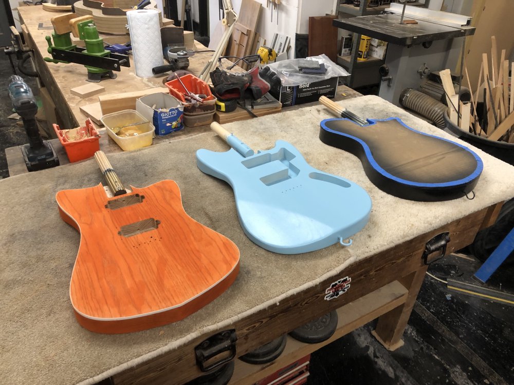 A photo of three guitar bodies sitting on a workbench. The first is a modernish offset shape with natural wood stained orange and a while plastic binding along the top edge. The second is a more classic offset painted sonic blue. The last is a rounded single cutaway design with black around the edges and a cardboard mask on top to protect the face from getting paint on it during spraying.