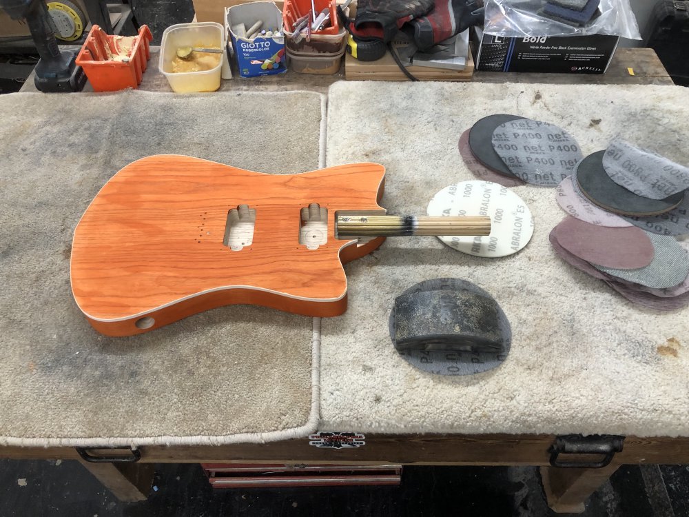 A photo of the orange-stained guitar body on the workbench, surrounded by sanding disks.