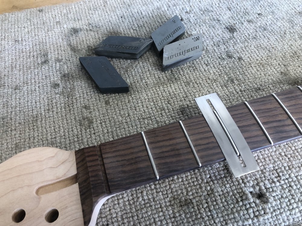 A photo of the same guitar neck again, this time showing the top five frets or so. On the third fret along sits a small metal plate that has a slot cut out for the fret but protects the surrounding fretboard. Next to the neck sit a bunch of small things that look like oversized pencil-erasers in different shades of grey, marked with C, M, F, and SF (course, medium, fine, super fine)