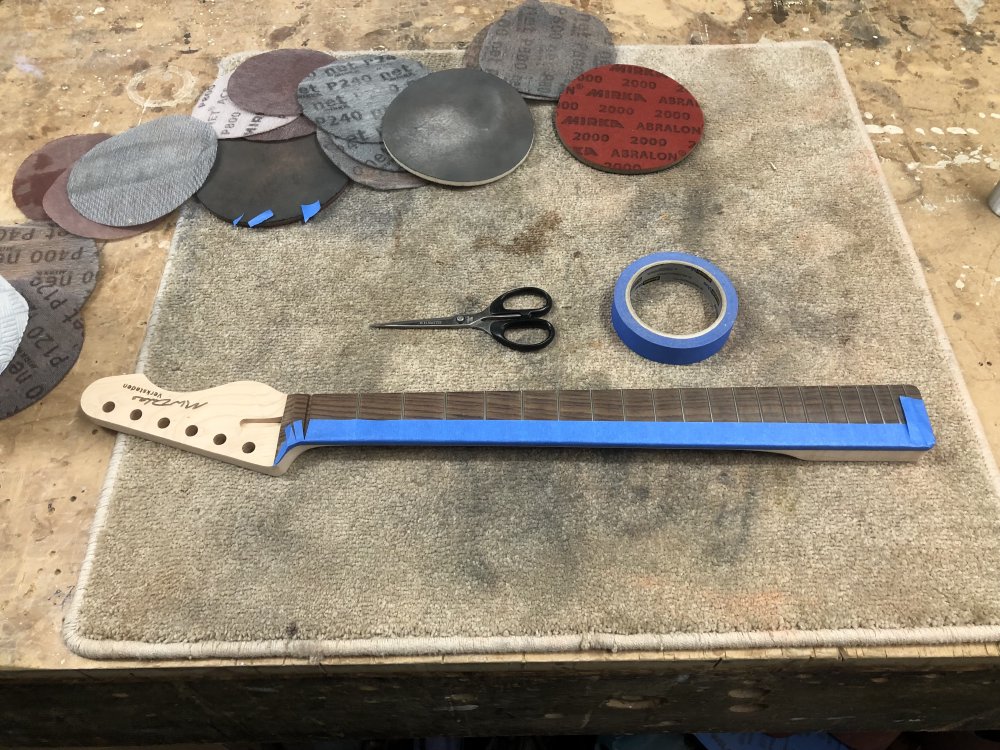 The guitar neck now sits on another workbench, surrounded by sanding disks, and the fretboard is half covered with masking tape, with the roll of tape and scissors sat poised for someone to finish the job.