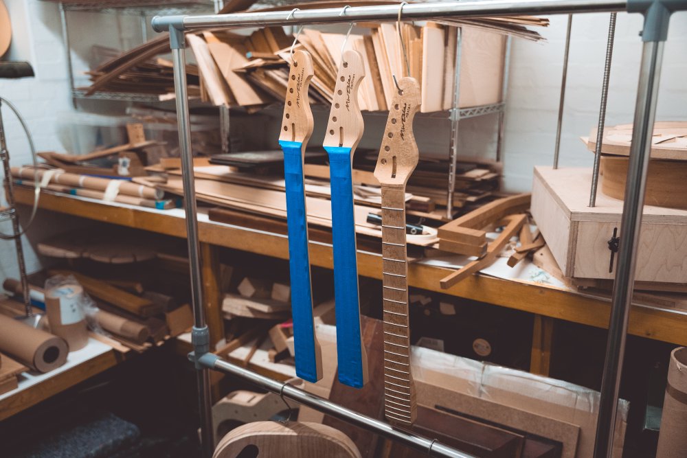 A photo of a trio of guitar necks hanging by a hook through a tuner hole, all on a rack in a room full of wood. All three are maple backs, and two have blue masking tape over the fretboards and one has a natural maple fretboard showing.