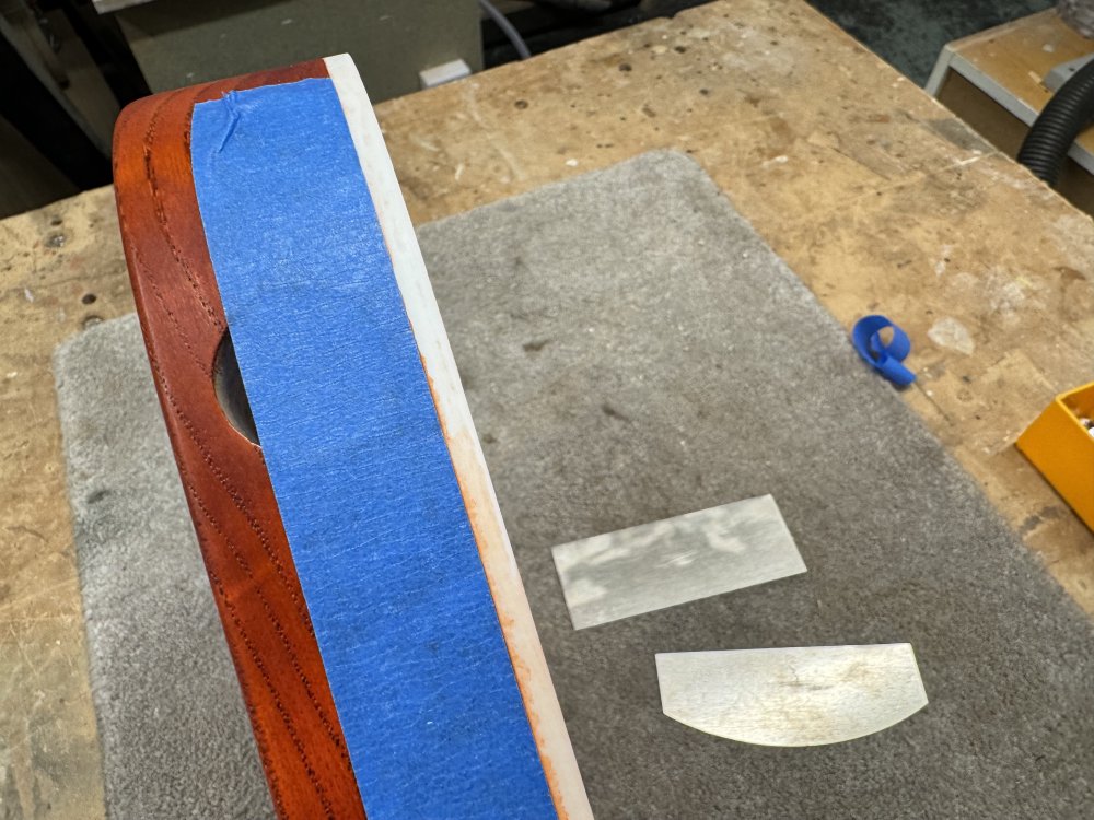 A photo of the side of the guitar body, where you can see the binding on the top edge, next to which is a strip of masking tape over the orange wood. Next to it on the bench are two metal wood scrapers.