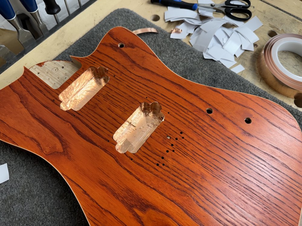 A shot of the orange stained guitar body in the workbench, and now the two pockets in the face for the pickups to go into are lined with copper foil tape. Next to the body is a lot of small bits of white backing paper showing it took a lot of tape to add to this guitar.
