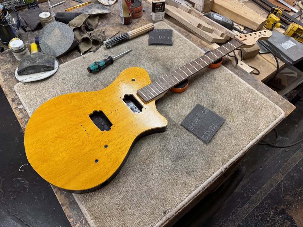 A photo of a work-in-progress guitar on the workbench, surrounded by tools and sanding pads. The guitar has a maple neck with a rosewood fretboard and 21 frets, and the body is a bright yellow stain on birds-eye maple on the front, with black and natural wood sides. There are holes in the body for components, but all are missing.
