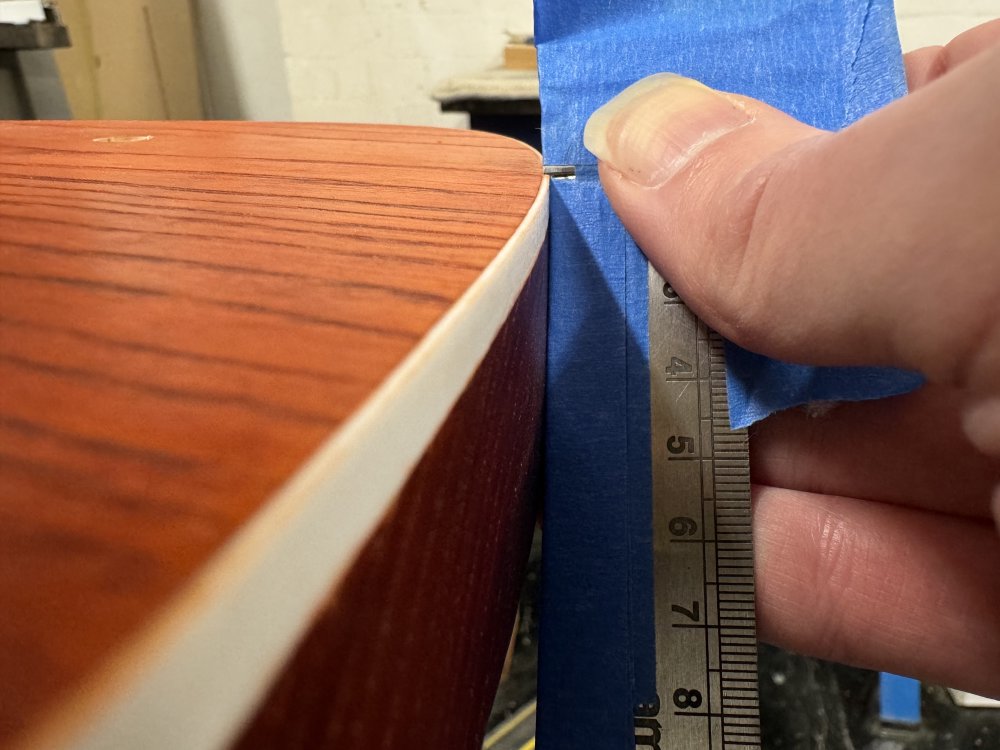A close up photo of the top edge of a guitar body that is stained orange and has white plastic binding around the top edge - the binding is 7mm deep down the side of the guitar but about 1mm wide into the top of the guitar. Presed up against the body is a metal ruler covered in masking tape, and out of which you can just make out the tiniest hint of a blade sticking out over the top thin edge of the binding.