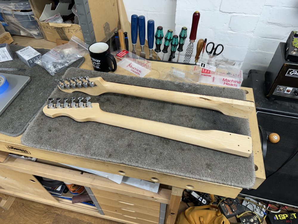 Two maple guitar necks sit on the workbench, rear showing, with tuners installed in their respective headstocks.