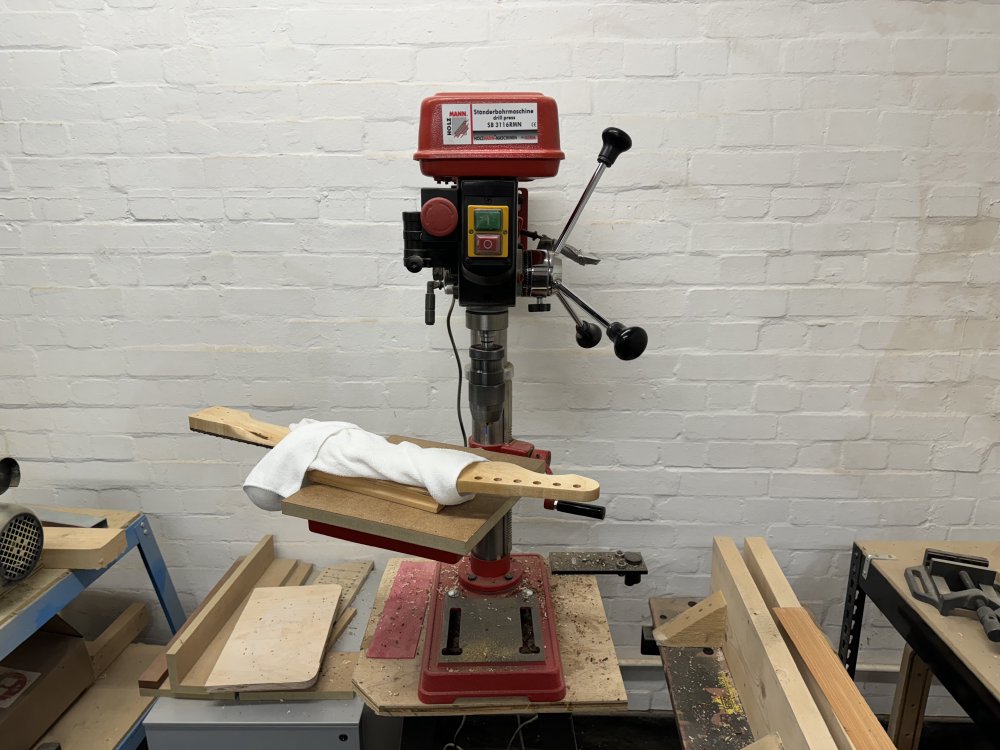 One of the guitar necks sits on the table of a pillar drill, wrapped in a towel to protect the fretboard as it's pressed into a radiusing block to hold the neck flat.