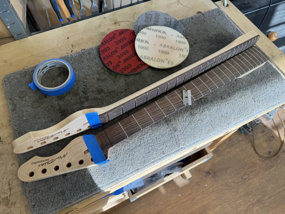 The two necks are now sat on a workbench fretboard side up (showing the rosewood fretboards). On top of one of the necks is a razor blade, and beside them is a pile of high-grit sanding disks.