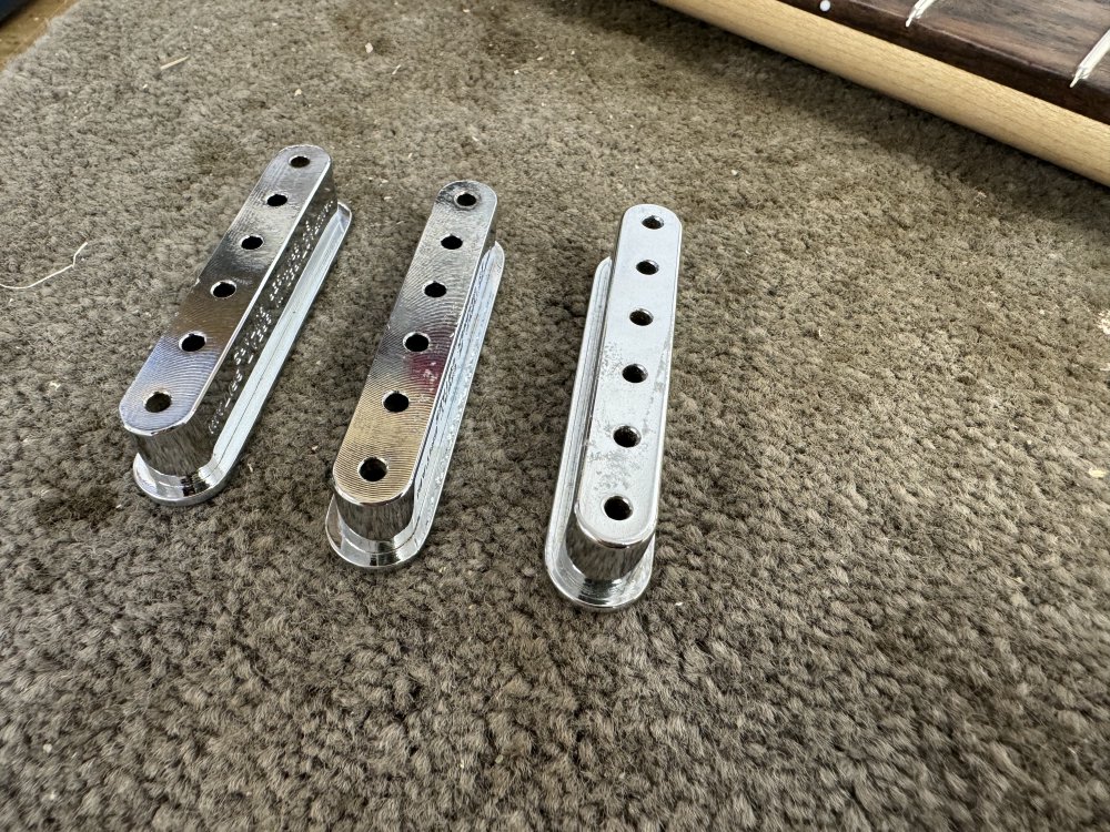 A picture of three metal string ferrule blocks - lozanges about 6 cm long with 6 holes in them for the strings to pass through and a lip on the top edge (currently at the bottom as they're sat upside down on the workbench. If you can look really close you can tell that two of them have been milled on teh bottom whereas the third one is smooth, indicating they were made differently despite otherwise being the same.