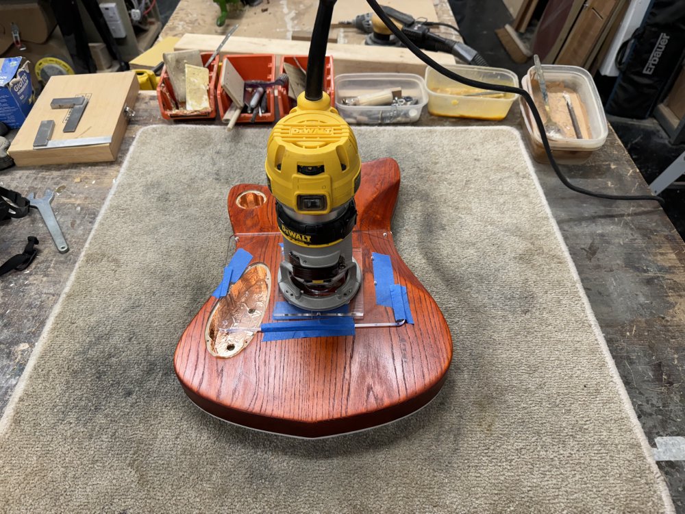 A photo of the nearly finished guitar body back on the workbench with a clear-acrylic template on the back of it stuck down with masking tape (what else?) and on that is sat a DeWalt palm router.