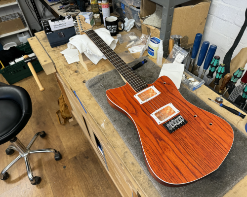 A mostly assembled guitar sits on the workbench! It is missing all the electronics, but otherwise looks like a guitar. Importantly it has strings on it! It has a vivid orange-stained wood body with white binding and white pickup mounts, and a maple neck with a rosewood fretboard.