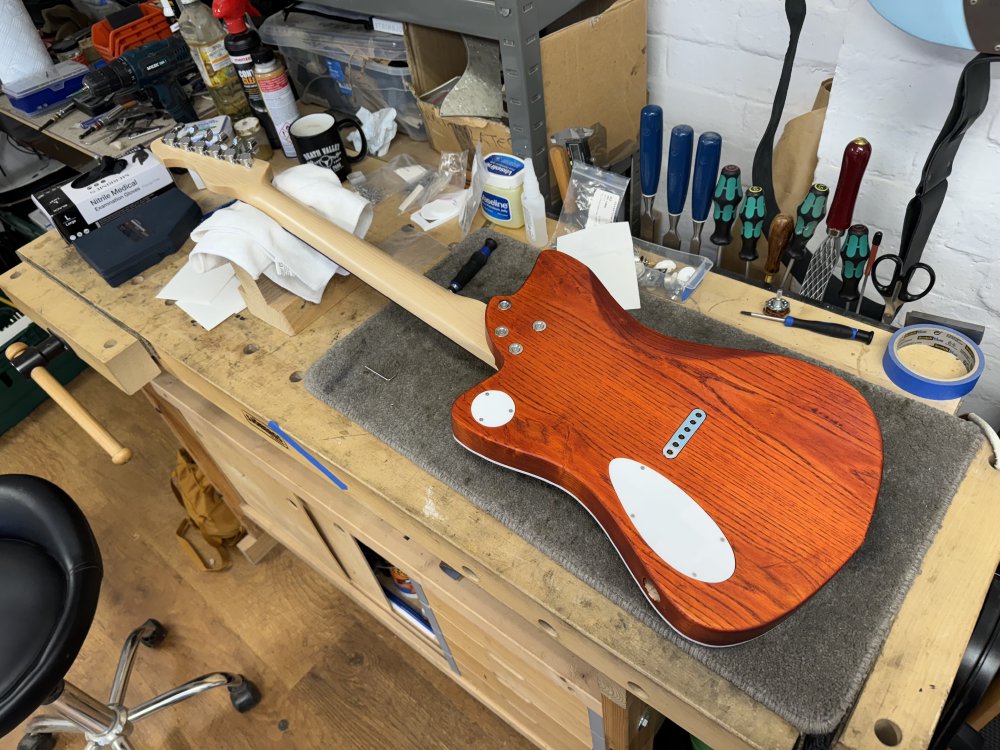 A rear view of the of the same guitar on the workbench. Here you can see that the neck is held on with four individual ferrules rather than the more traditional neck plate, we have the single string-ferrule block rather than individual string ferrules, and we have the two white acrylic covers in place that match the binding.