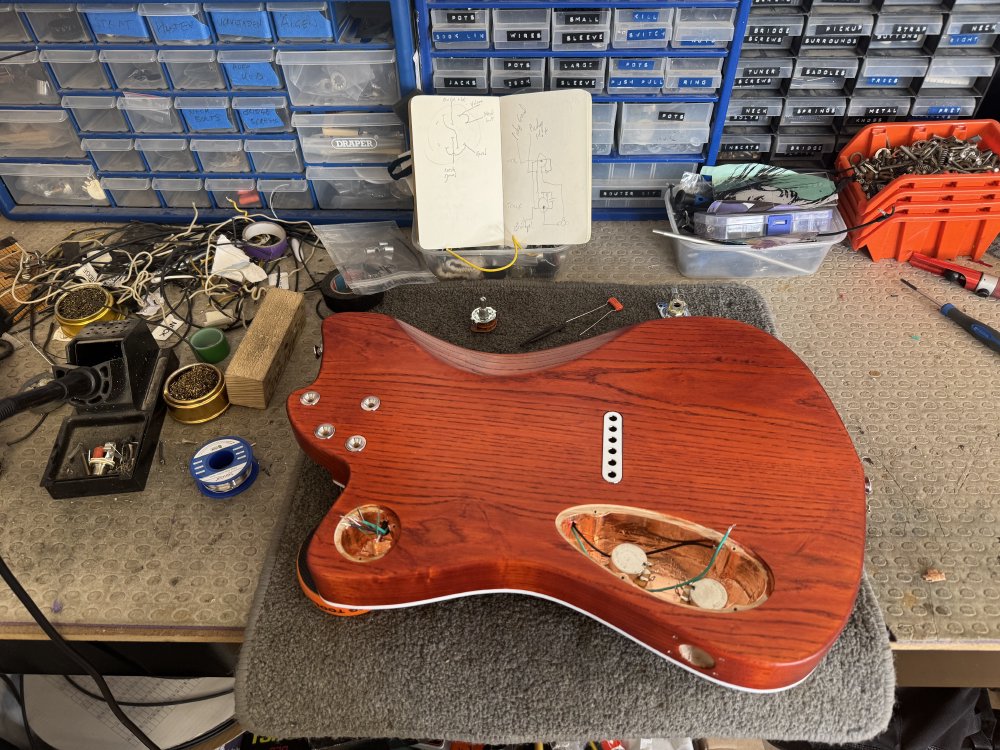 A photo of the back of the guitar body, now sat on an electronics workbench with soldering station to one side and electronics components scattered around.