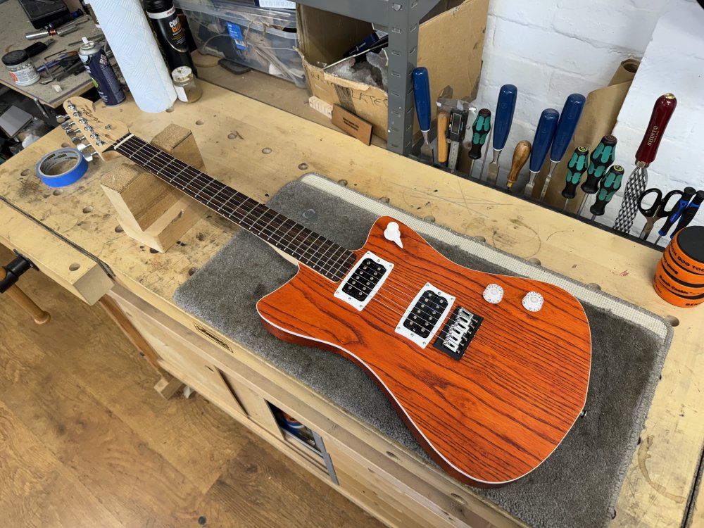 A photo of a guitar sat on a workshop bench. The guitar is an offset style guitar, with an orange stained natural wood finish, with white plastic binding around the edge. It has two humbucker pickups, and a maple neck with a rosewood fretboard.
