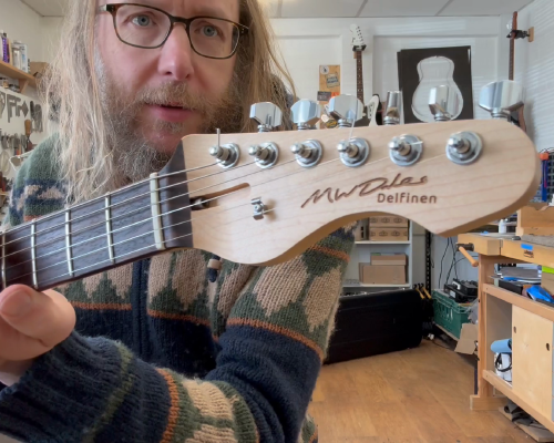 A still from a video showing Michael sat in front of the camera holding a guitar, showing the headstock of it to the camera. On the headstock it says "MWDales" in script and "Delfinen" under that.