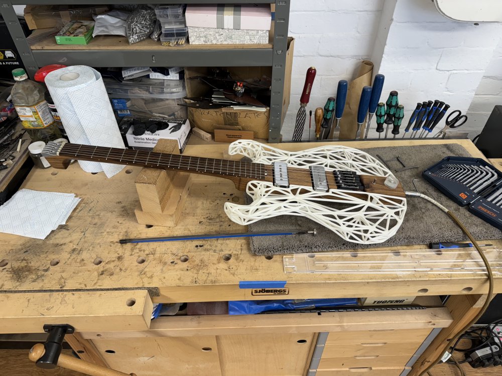 A photo of an unusual guitar on a workbench with some tools and a truss-rod beside it. The guitar is unusual as the sides are made fo white 3D-printed plastic and there is no headstock.
