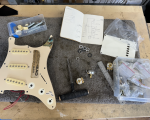 A photo of a pickguard from a stratocaster style guitar on a workbench surrounded by electronics parts and a notebook with a wiring diagram hand draw in it.