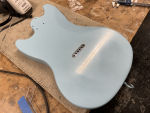 A photo of a blue-painted mustang-style guitar body is sat on the workbench showing a string-ferrule block in place on the rear.