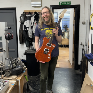 A photo of a bearded luthier standing in a workshop holding an orange offset electric guitar.